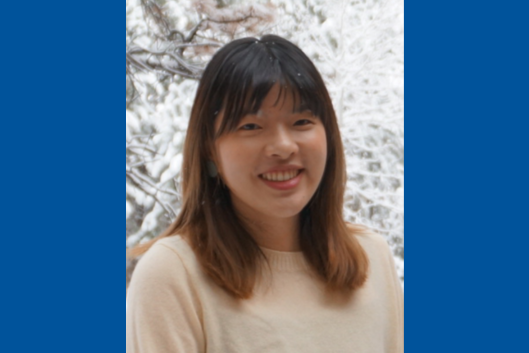 A photo of Tiffany Tu, a young woman with shoulder-length dark brown hair and bangs. She is smiling at the camera. She is wearing a cream-colored sweater and standing outdoors with snow-covered trees in the background.